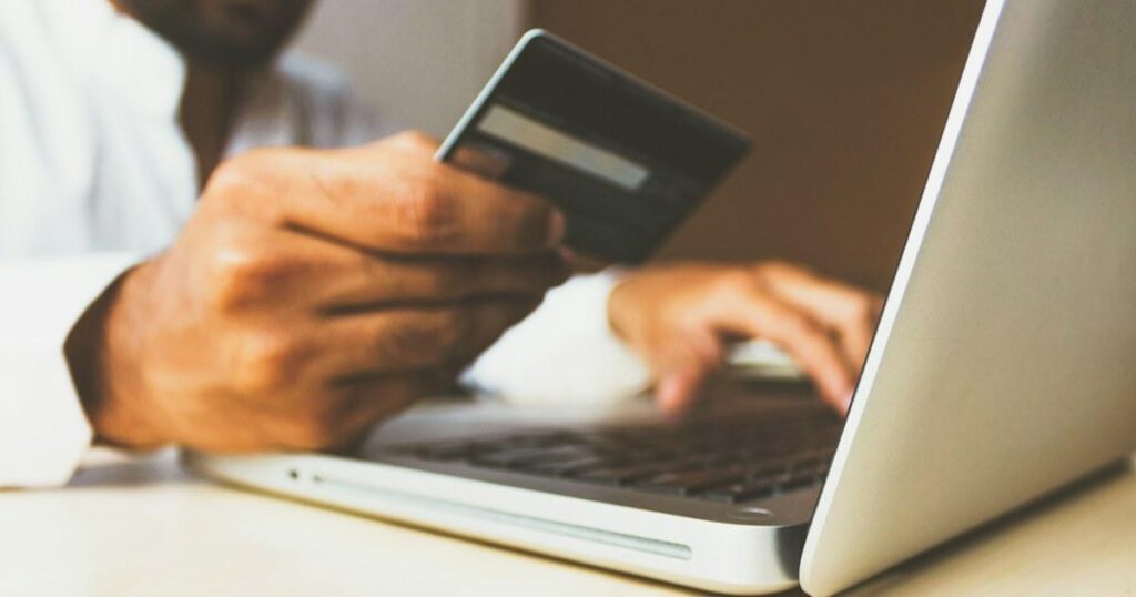 Person holding a payment card in one hand while typing on a silver laptop keyboard at a white desk